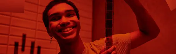 Веселий афроамериканський чоловік, що висить свіжо розвинену плівкову стрічку в червоно-світлій темній кімнаті, банер — стокове фото