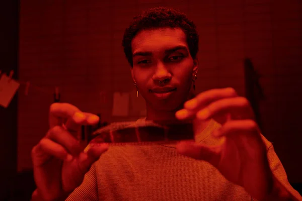 Satisfecho afroamericano fotógrafo examinar desarrollado tira de película en un rojo-iluminado cuarto oscuro - foto de stock