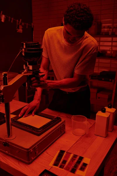 Photographe à la peau foncée se concentre sur le processus délicat d'agrandissement du film dans la chambre noire avec lumière rouge — Photo de stock