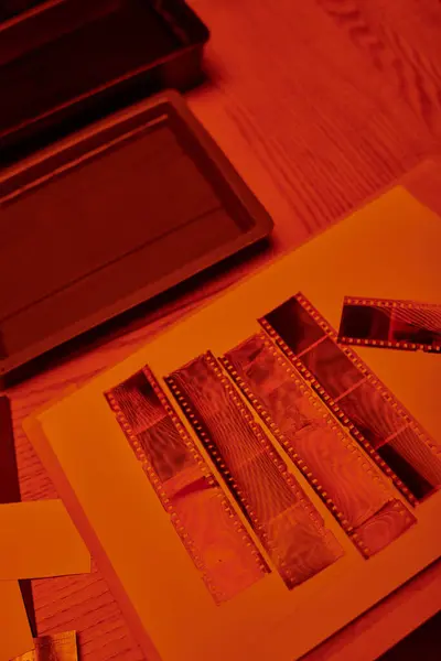 Tiras de película desarrolladas en una mesa junto al equipo de fotografía del cuarto oscuro, con luz roja de seguridad - foto de stock