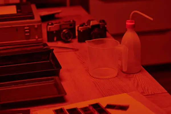 A table with analog camera and measuring cup for film development in darkroom with red light — Stock Photo