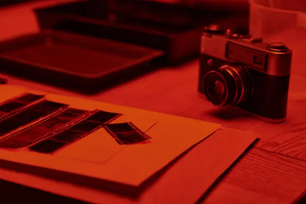 Стіл з аналоговою камерою і смужками плівки під сяйвом червоного світла в темній кімнаті, поза часом — стокове фото