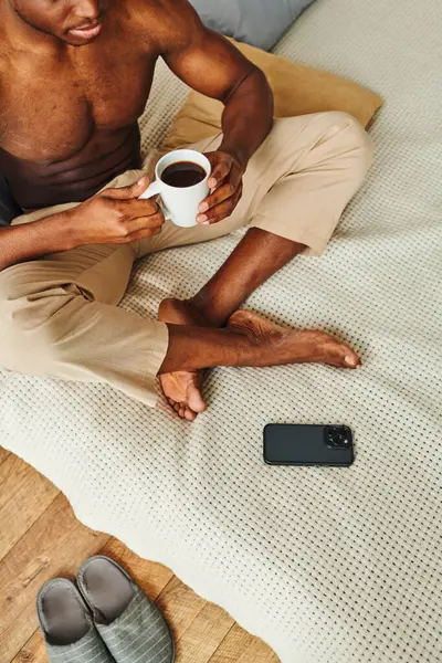 Африканский американец в пижамных штанах сидит на кровати с кофейной чашкой рядом со смартфоном, обрезанный выстрел — стоковое фото