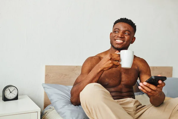 Feliz sem camisa afro-americano homem com xícara de café e smartphone sentado e olhando para a cama — Fotografia de Stock