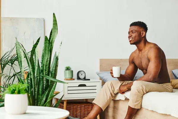 Усміхнений без сорочки афроамериканський чоловік з чашкою кави сидить біля зелених горщиків рослин у спальні — стокове фото