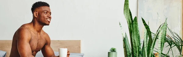 Sonriente afroamericano sin camisa hombre sentado con taza de café cerca de plantas verdes en el dormitorio, pancarta - foto de stock
