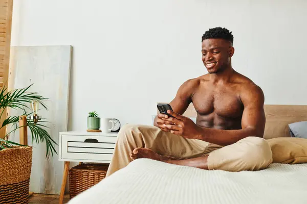 Радісний і м'язистий афроамериканський американський в піжамі штани, що переглядають соціальні медіа om смартфон у спальні — стокове фото