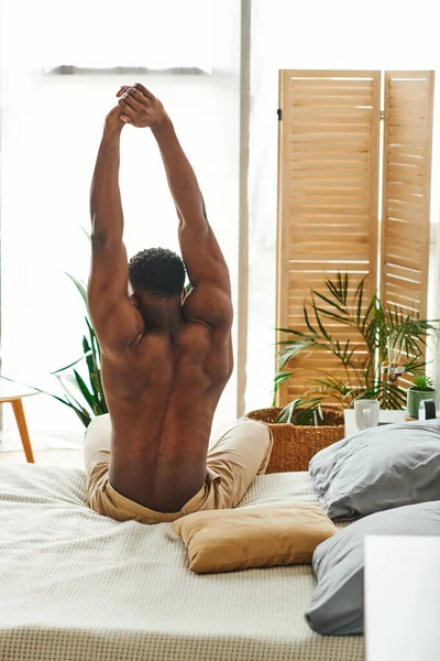 Без сорочки і сильний афроамериканський хлопець сидить і тягнеться на ліжку вранці, вид ззаду — стокове фото