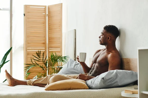 Homme afro-américain torse nu rêveur avec tasse de café assis sur le lit et regardant loin le matin — Photo de stock