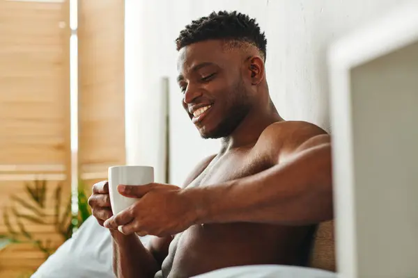 Erfreut afrikanisch-amerikanischer Mann mit hemdlosem Oberkörper sitzt mit Morgenkaffee im modernen Schlafzimmer — Stockfoto