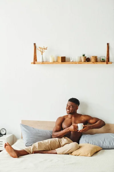 Cheerful athletic african american man in pajama pants sitting with coffee cup on bed in morning — Stock Photo