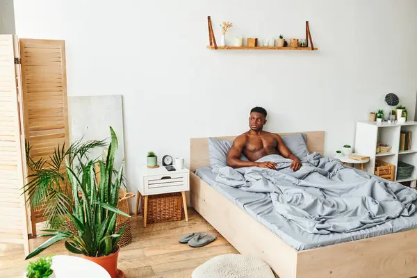 Shirtless muscular african american man sitting on bed in spacious bedroom with green potted plants — Stock Photo