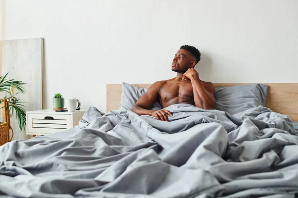 Jeune homme afro-américain torse nu assis sur une literie grise et regardant loin dans la chambre moderne — Photo de stock