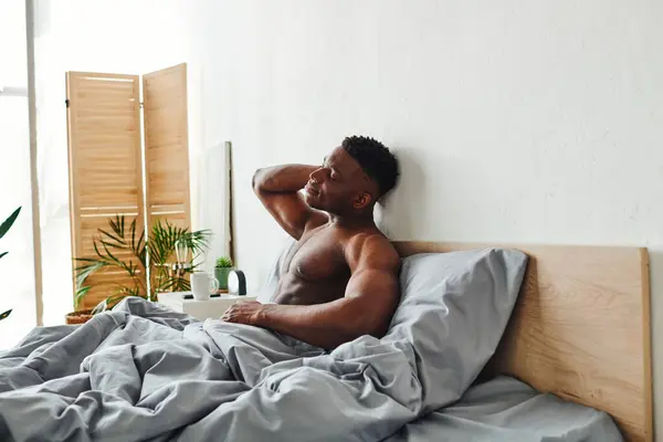 Homem americano africano satisfeito com corpo muscular sem camisa e olhos fechados sentados na cama de manhã — Fotografia de Stock