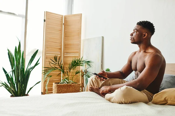 Verträumter afrikanisch-amerikanischer Mann in Pyjamahose sitzt mit Smartphone im Bett und schaut weg — Stockfoto
