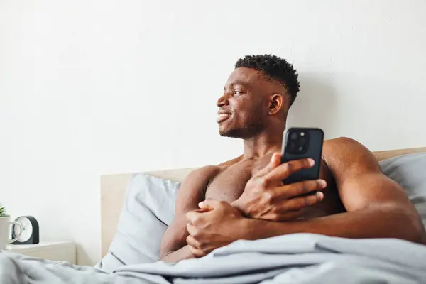 Fröhlich muskulöser Afroamerikaner mit Handy schaut im modernen Schlafzimmer auf gemütlichem Bett weg — Stockfoto
