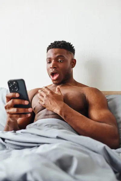 Amazed muscular african american man looking at smartphone while browsing internet in bedroom — Stock Photo