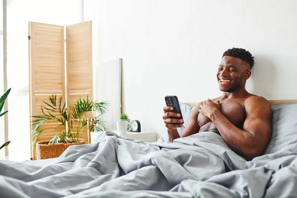 Ridere uomo afroamericano senza maglietta guardando smartphone durante la navigazione in internet in camera da letto — Foto stock