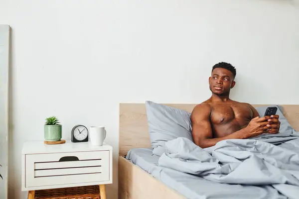 Thoughtful african american man with shirtless muscular body and smartphone looking away in bedroom — Stock Photo