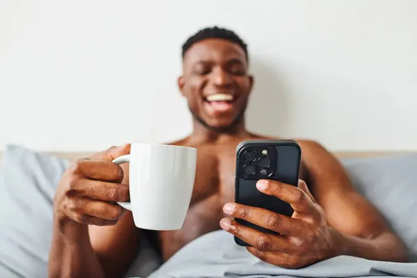 Emocionado hombre afroamericano sin camisa con café de la mañana y teléfono móvil riendo en el dormitorio - foto de stock