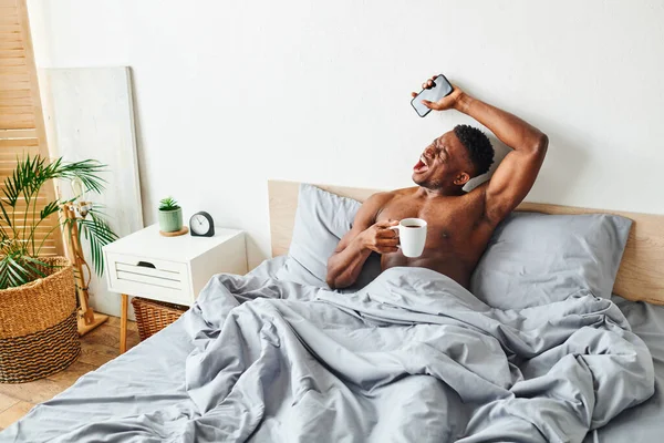 Молодой африканский американец с чашкой кофе и смартфоном зевает и растягивается просыпаясь на кровати — стоковое фото