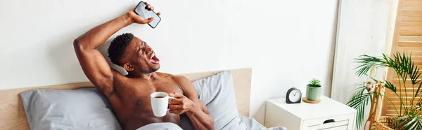 Uomo afroamericano muscoloso con tazza di caffè e smartphone sbadigliando e allungando sul letto, banner — Foto stock