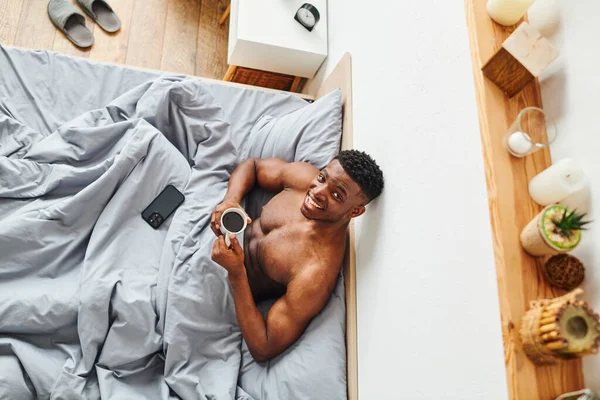 Vista dall'alto di allegro uomo africano americano senza maglietta con caffè mattutino sorridente alla macchina fotografica sul letto — Foto stock