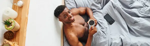 Vista superior do homem americano africano muscular feliz com café da manhã olhando para a câmera na cama, banner — Fotografia de Stock