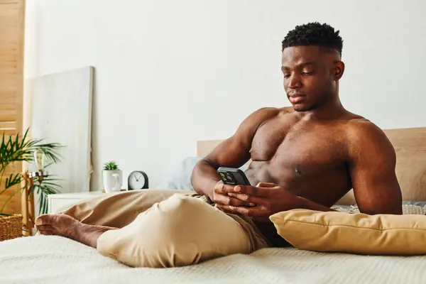 Athletic african american man in pajama pants messaging on mobile phone on cozy bed in morning — Stock Photo