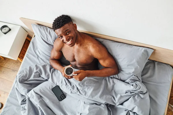 Top view of carefree athletic african american man with morning coffee looking at camera on bed — Stock Photo