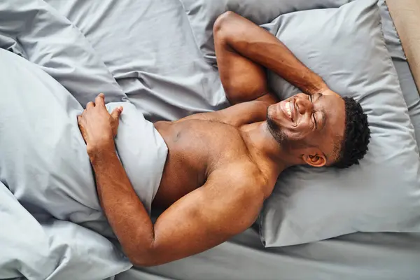 Top view of happy muscular african american man dreaming and smiling on grey comfortable bedding — Stock Photo