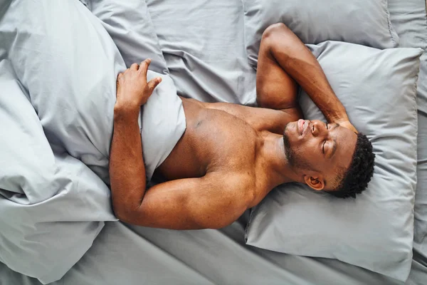 Von oben: Afrikanisch-amerikanischer Mann mit muskulösem Körper schläft auf grauem, bequemen Bettzeug zu Hause — Stockfoto