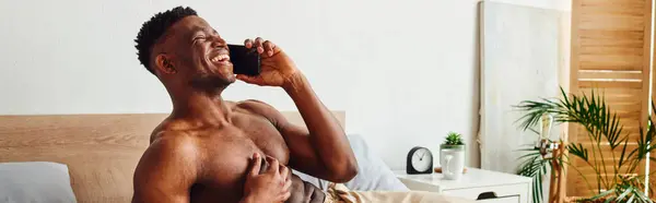 Fröhlicher muskulöser afrikanisch-amerikanischer Mann in Pyjamahose, der auf dem Smartphone im gemütlichen Schlafzimmer spricht, Banner — Stockfoto