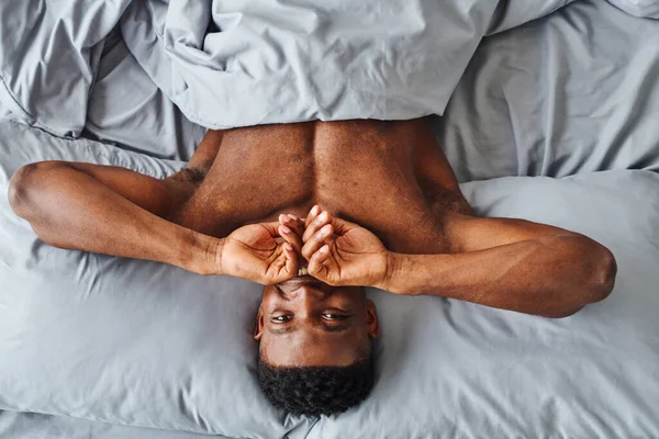 Draufsicht des fröhlichen afrikanisch-amerikanischen Mannes, der den Mund bedeckt und in die Kamera schaut, die auf dem Bett aufwacht — Stockfoto