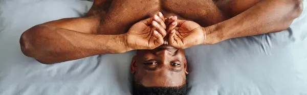 Joyful african american guy covering mouth and smiling at camera waking up on bed, top view, banner — Stock Photo