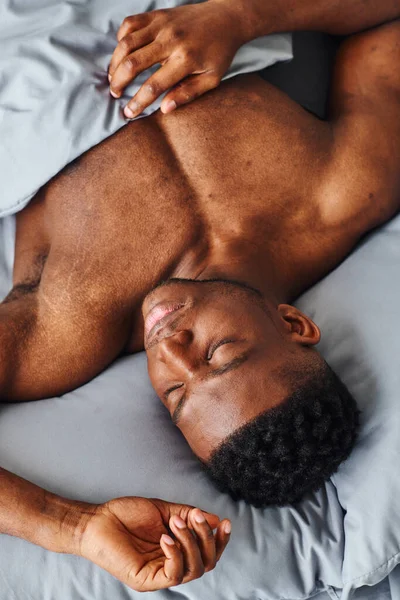 Draufsicht auf einen jungen afrikanisch-amerikanischen Mann mit muskulösem Körper, der morgens auf grauem Bettzeug schläft — Stockfoto