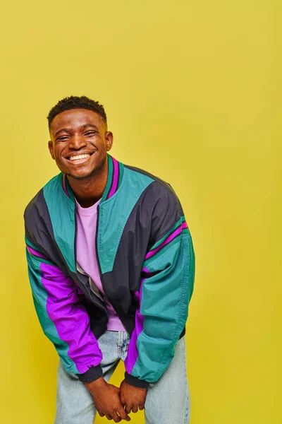 Overjoyed trendy african american man in bright windbreaker smiling and posing on yellow backdrop — Stock Photo