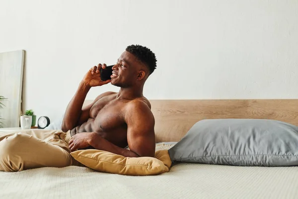 Feliz sem camisa afro-americano homem em calças de pijama falando no smartphone perto de travesseiros na cama acolhedora — Fotografia de Stock