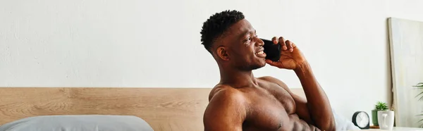 Lächelnder muskulöser afrikanisch-amerikanischer Mann, der im modernen gemütlichen Schlafzimmer auf dem Handy spricht, Banner — Stockfoto