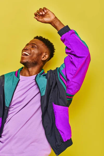 Hombre afroamericano lleno de alegría en chaqueta cortavientos colorido agitando la mano y mirando hacia otro lado en amarillo - foto de stock