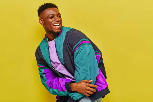 Excited african american man in bright windbreaker jacket laughing and looking away on yellow — Stock Photo