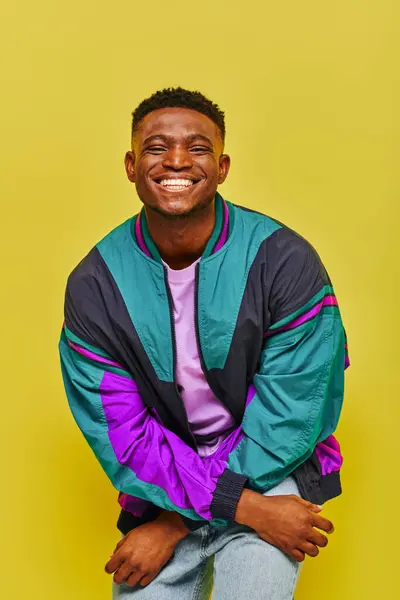 Overjoyed and stylish african american man in bright windbreaker jacket looking at camera on yellow — Stock Photo