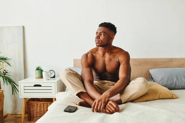 Uomo africano muscoloso premuroso in pigiama pantaloni seduto sul letto vicino smartphone e guardando altrove — Foto stock