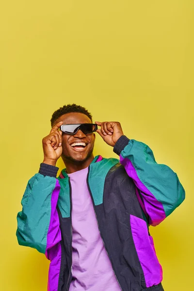 Joyful african american man in colorful windbreaker jacket adjusting trendy sunglasses on yellow — Stock Photo