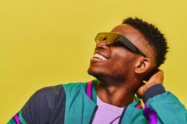 Homme afro-américain insouciant dans une veste coupe-vent et des lunettes de soleil élégantes regardant loin sur jaune — Photo de stock