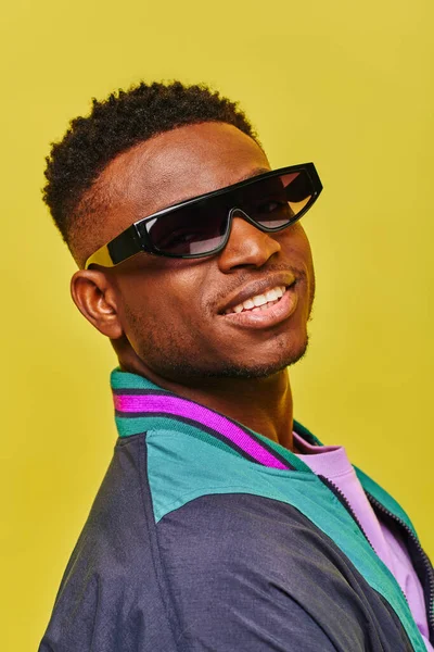 Portrait of african american man in sunglasses and colorful jacket smiling at camera on yellow — Stock Photo