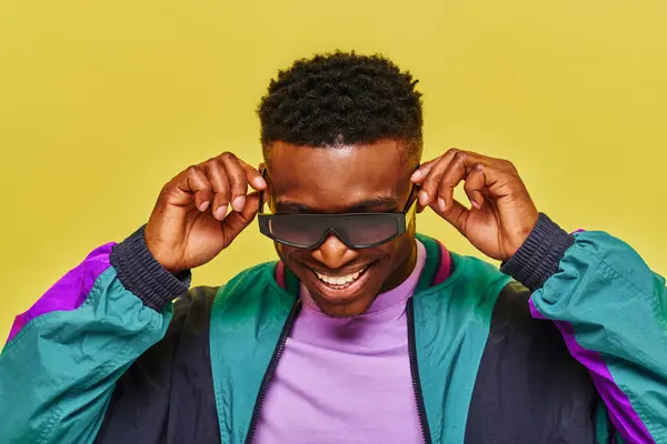 Elegante uomo africano in giacca colorata regolazione occhiali da sole e sorridente su sfondo giallo — Foto stock