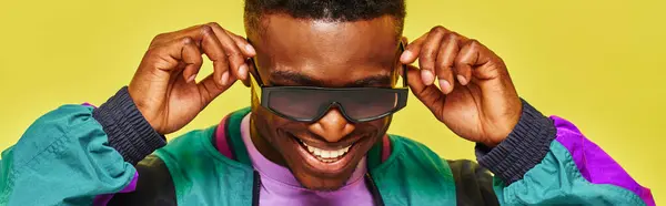 Trendy african guy in bright jacket adjusting sunglasses and smiling on yellow backdrop, banner — Stock Photo