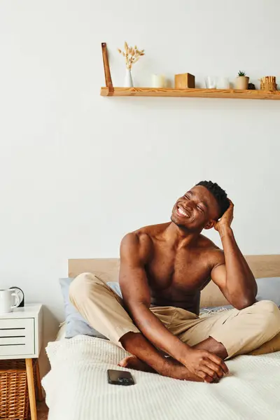 Riant sans chemise homme afro-américain en pyjama pantalon regardant loin près du smartphone sur le lit à la maison — Photo de stock