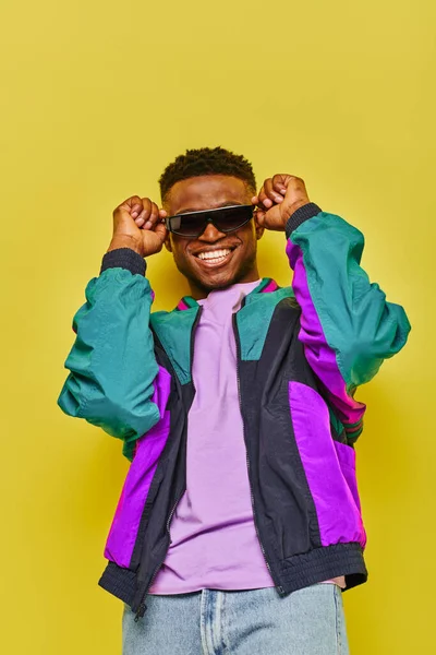 Homme afro-américain ravi en veste lumineuse et des lunettes de soleil à la mode souriant à la caméra sur jaune — Photo de stock